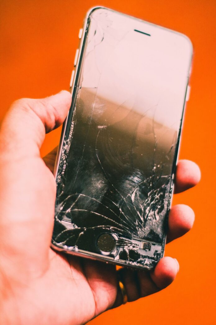 hardik-sharma-27mgWH0pzhA-unsplash (1) Defektes Smartphone mit stark gesplittertem Displayglas wird in einer Hand vor orangefarbenem Hintergrund gehalten