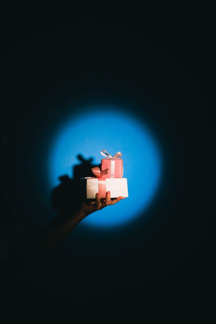 pexels-shvetsa-6631417 (1) Hand hält zwei rosa-weiße Geschenkboxen im Spotlicht vor blauem Hintergrund